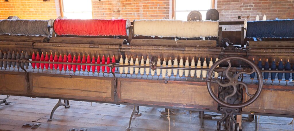Machine containing gray, red, white, and blue rollers with woolen fiber and smaller spindles the yarn.