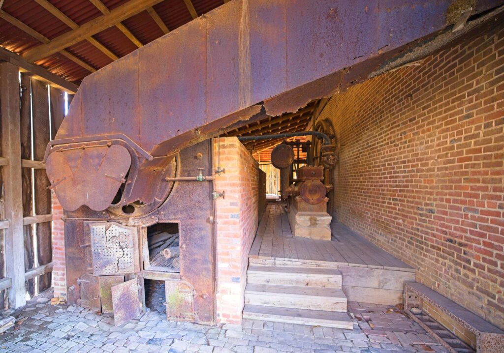 A mid-19th century boiler on the left and a steam engine on the right sitting against a brick wall.